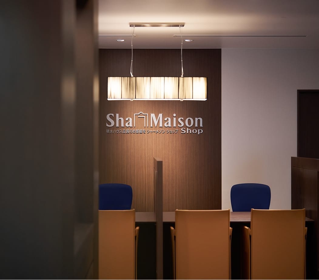 積水ハウス品質のお部屋を探せる「ShaMaison Shop（シャーメゾンショップ）」の受付カウンター。木目調の壁面にブランドロゴとペンダントライトが設置され、手前にはオレンジ色の椅子とパソコンモニターが並ぶ落ち着いた雰囲気の店内。